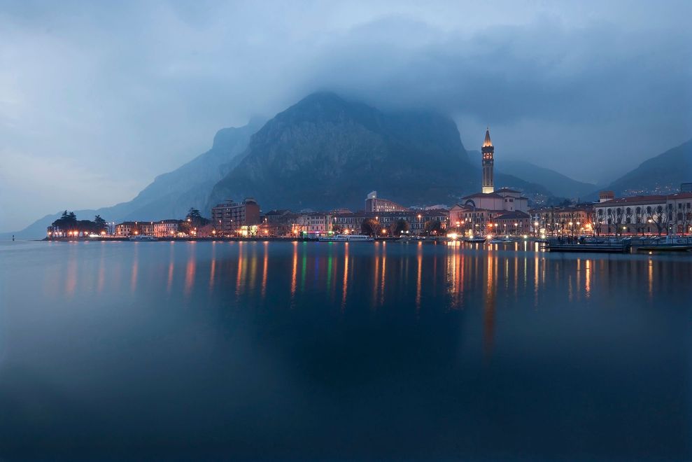 Cittadina lacustre avvolta nella nebbia al crepuscolo, con le montagne sullo sfondo e i riflessi luminosi sull'acqua.