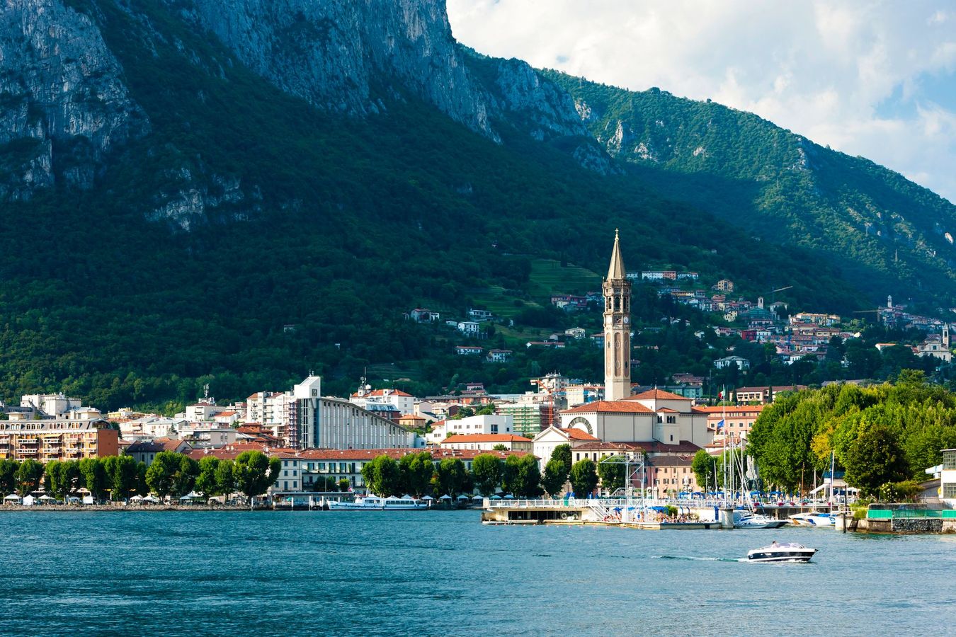 Cittadina lacustre con campanile, montagne sullo sfondo e barche sull'acqua blu.