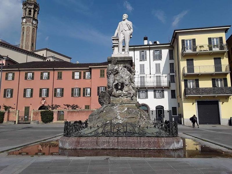 Statua su un piedistallo in una piazza soleggiata di una città europea, con edifici colorati e una torre sullo sfondo.