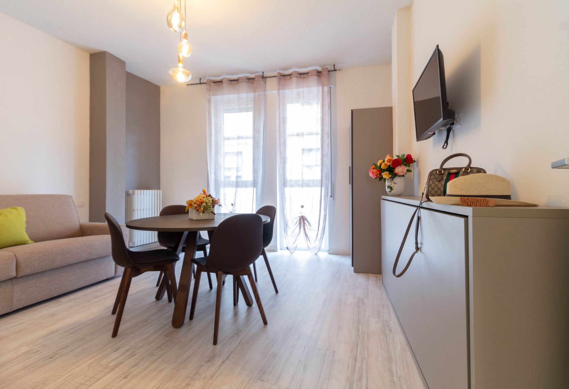 Luminosa sala da pranzo-soggiorno moderna con tavolo rotondo, divano grigio, TV a parete e tende trasparenti vicino alla porta del balcone.