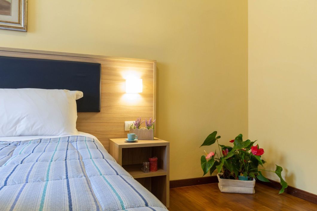 Piccola camera da letto con letto, comodino in legno, lampada da parete e una pianta in vaso sul pavimento.