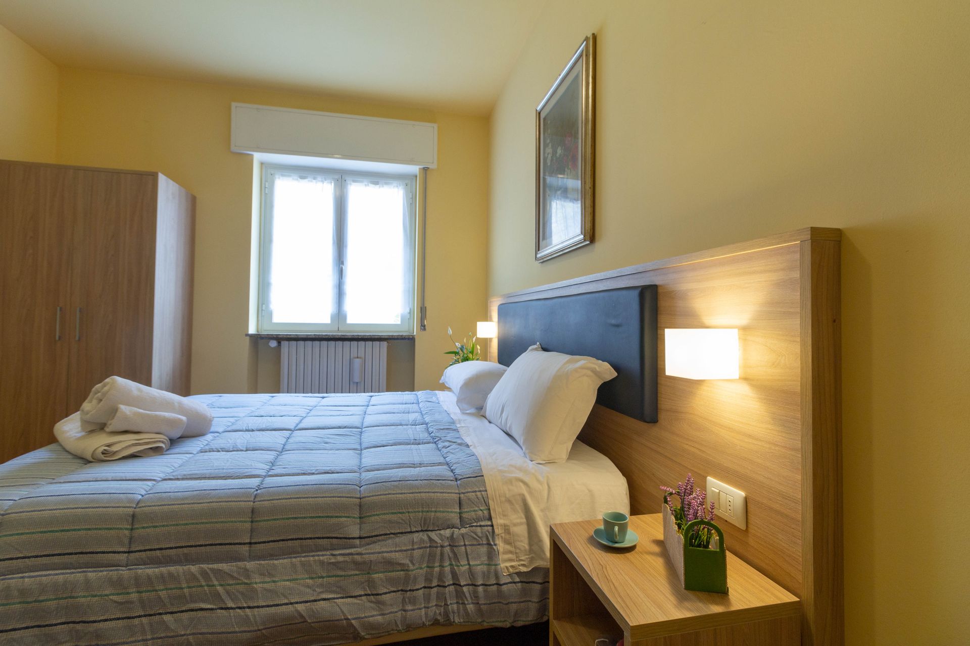 Camera d'albergo pulita e ordinata, con letto matrimoniale, lampada da comodino, finestra e mobili in legno.
