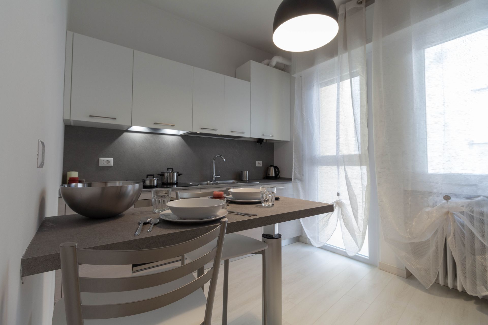 Cucina e zona pranzo moderne con mobili bianchi, tavolo in legno e tende trasparenti accanto a una finestra luminosa.