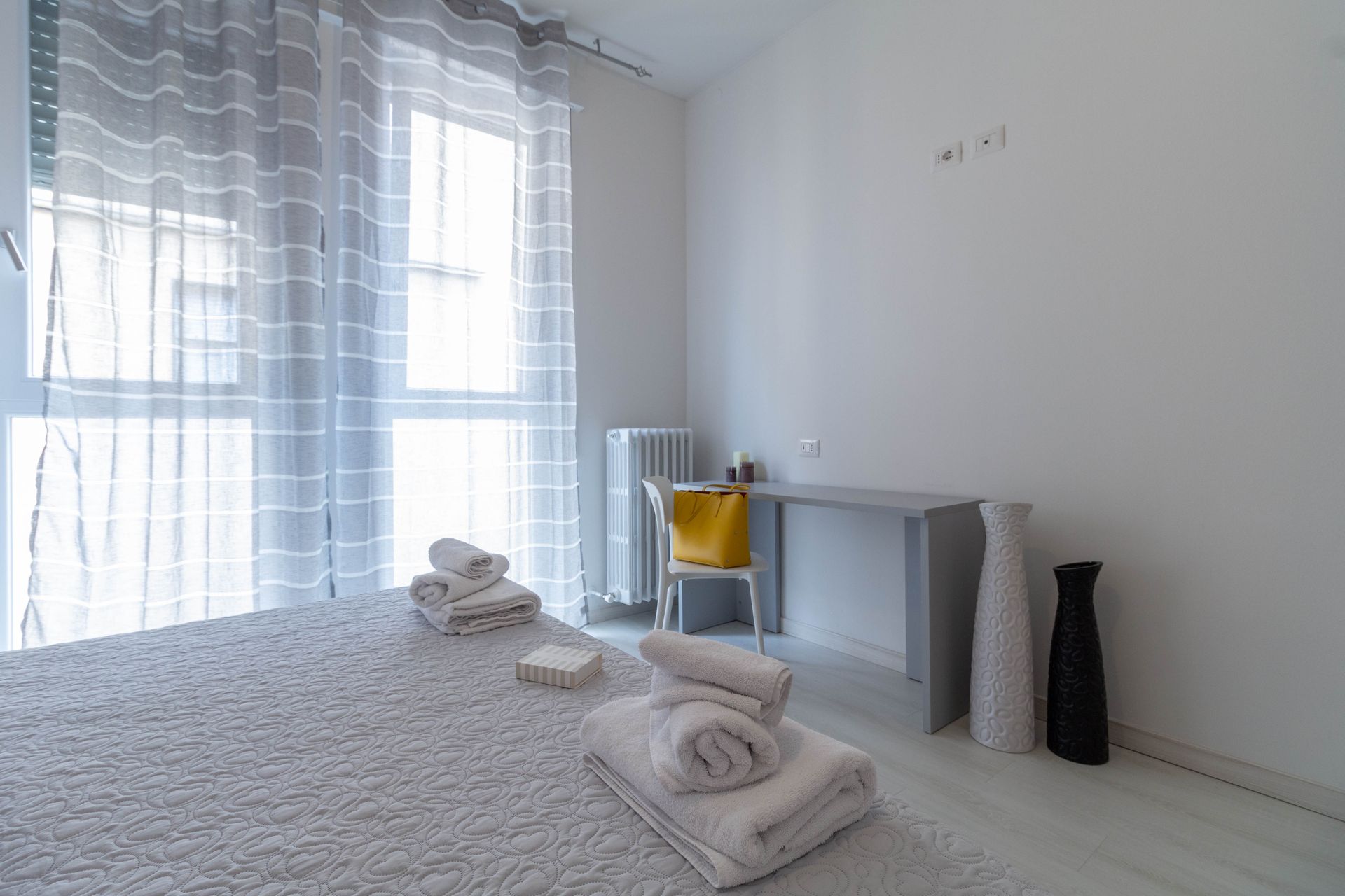 Luminosa stanza in stile minimalista con due lettini da massaggio, asciugamani piegati, una scrivania, una sedia e ampie finestre con tende trasparenti.