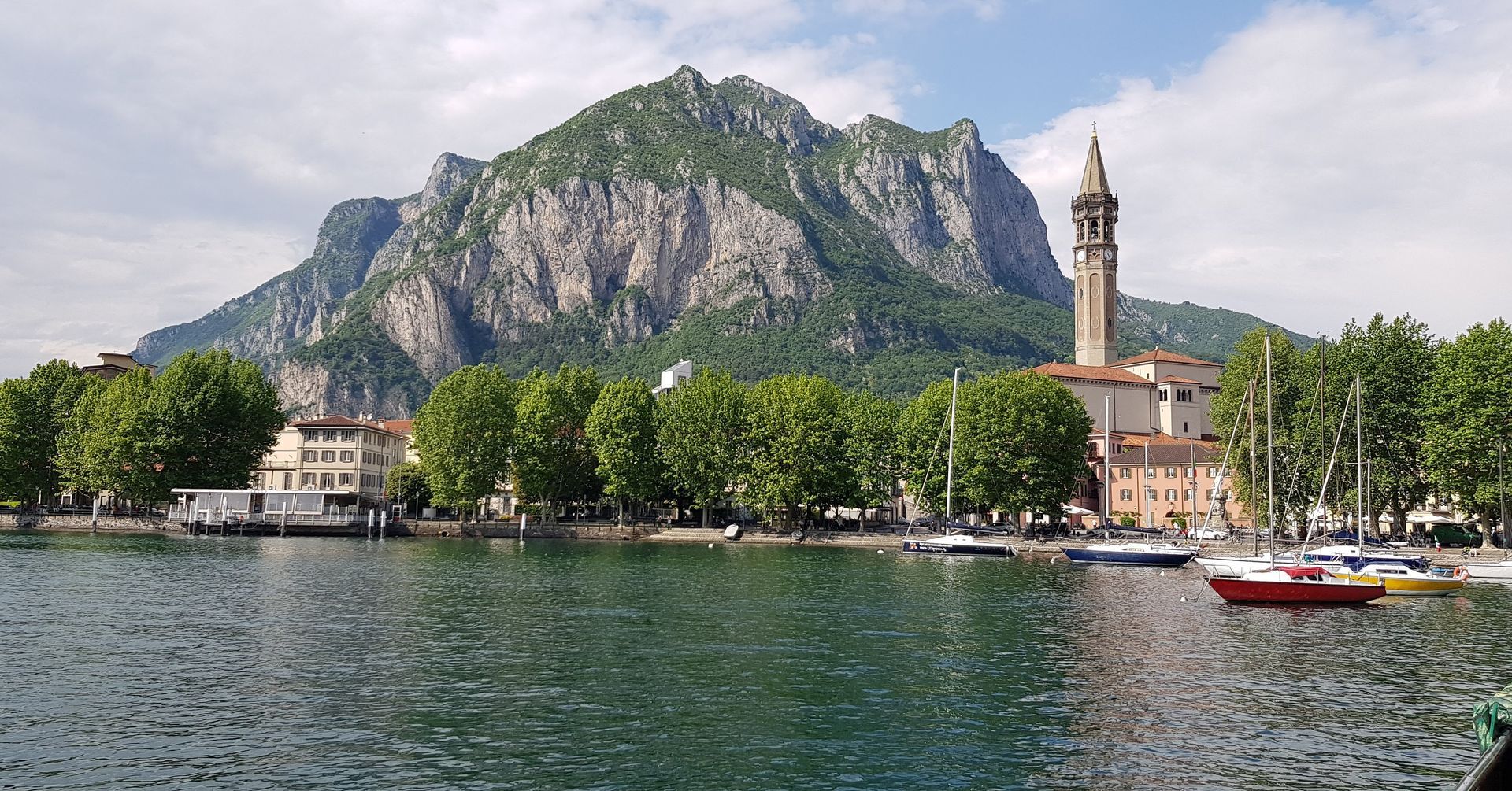 Cittadina lacustre con una montagna rocciosa, un campanile, alberi e una piccola barca su acque calme.
