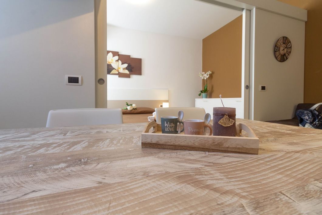 Luminoso soggiorno-sala da pranzo con divano beige, tavolo rotondo, sedie scure e tende trasparenti vicino a una finestra