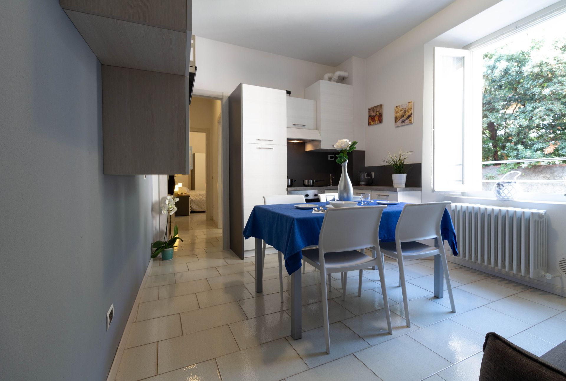 Luminosa cucina-sala da pranzo con tovaglia blu, sedie, pavimento piastrellato e luce naturale proveniente da un'ampia finestra.