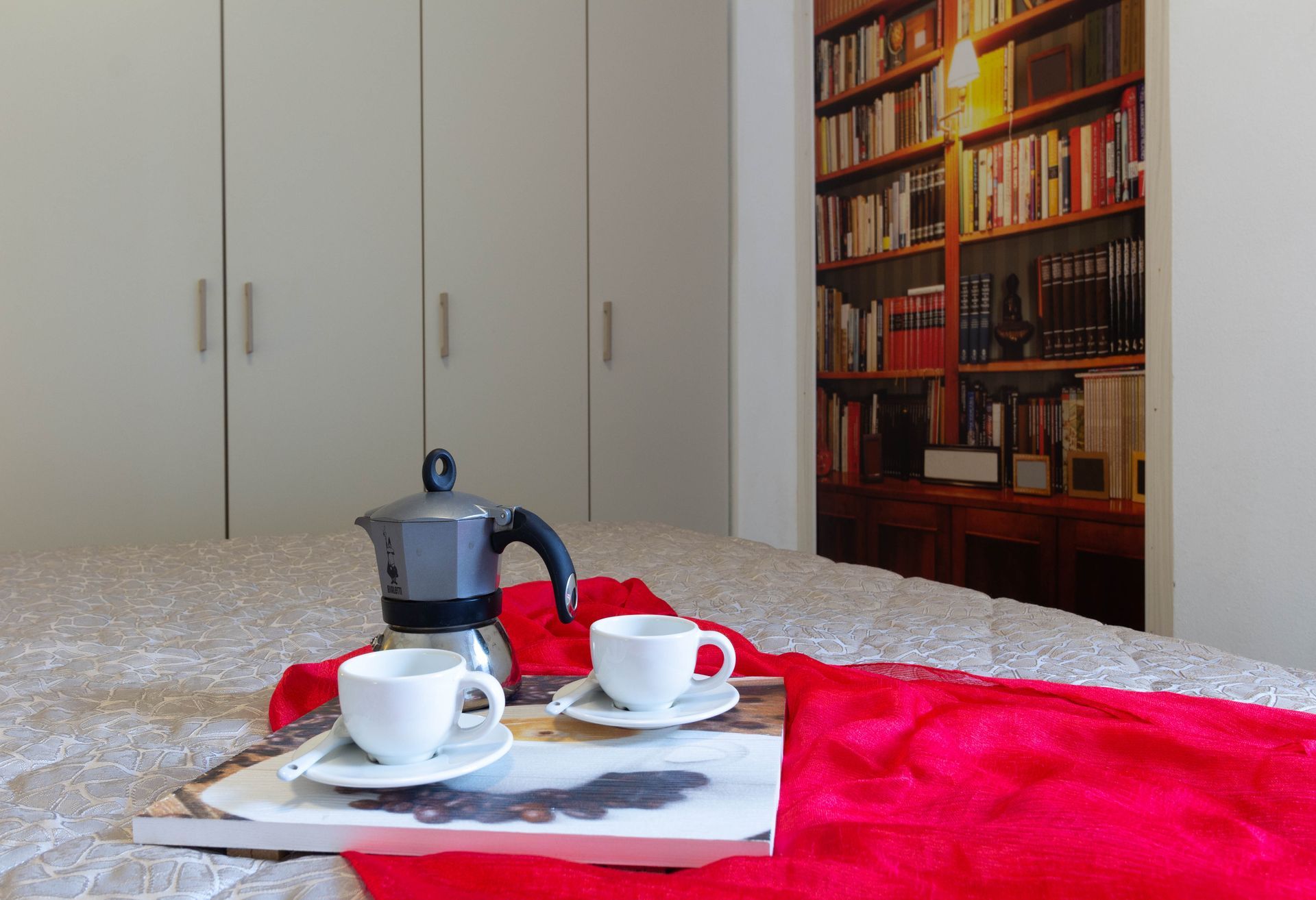 Tazze da caffè e una moka su un vassoio sopra un letto con una coperta rossa in una camera da letto.