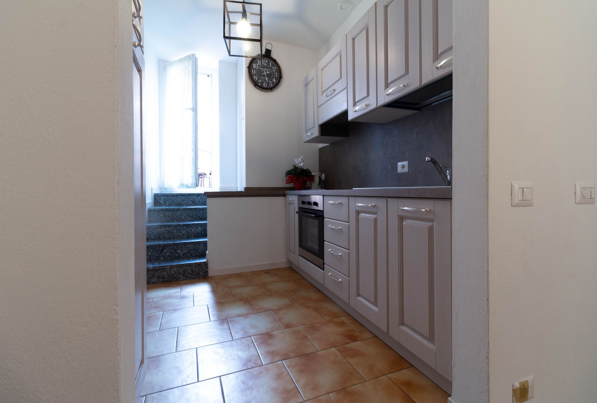 Cucina stretta con mobili grigi, pavimento piastrellato e scala che conduce a una porta luminosa.
