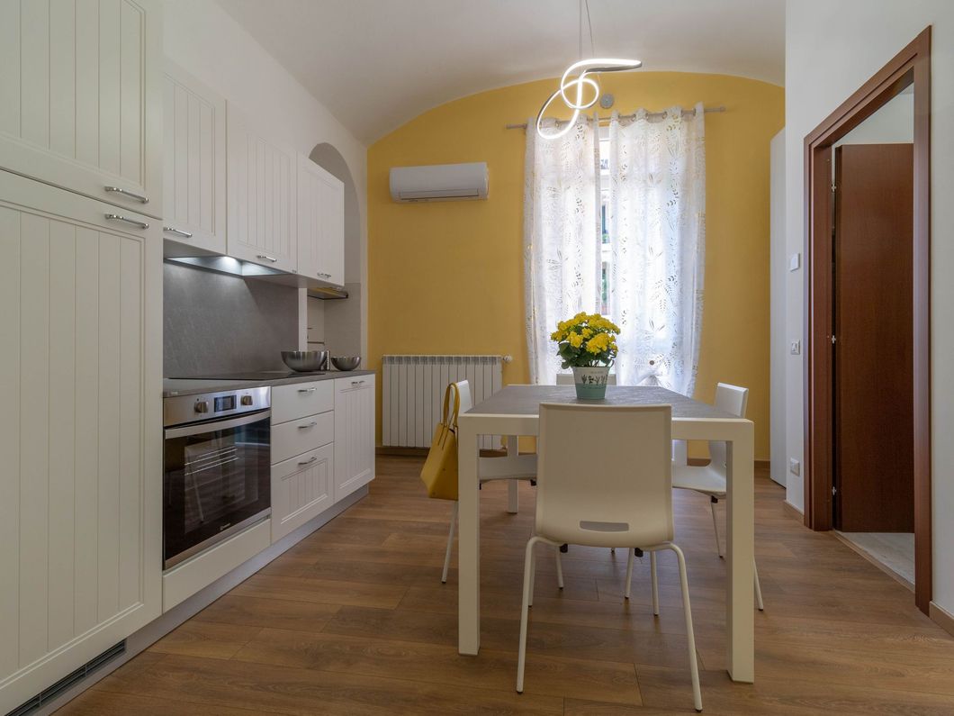Cucina luminosa con mobili bianchi, pavimento in legno, tavolo da pranzo e un vaso di fiori gialli accanto a una finestra soleggiata.