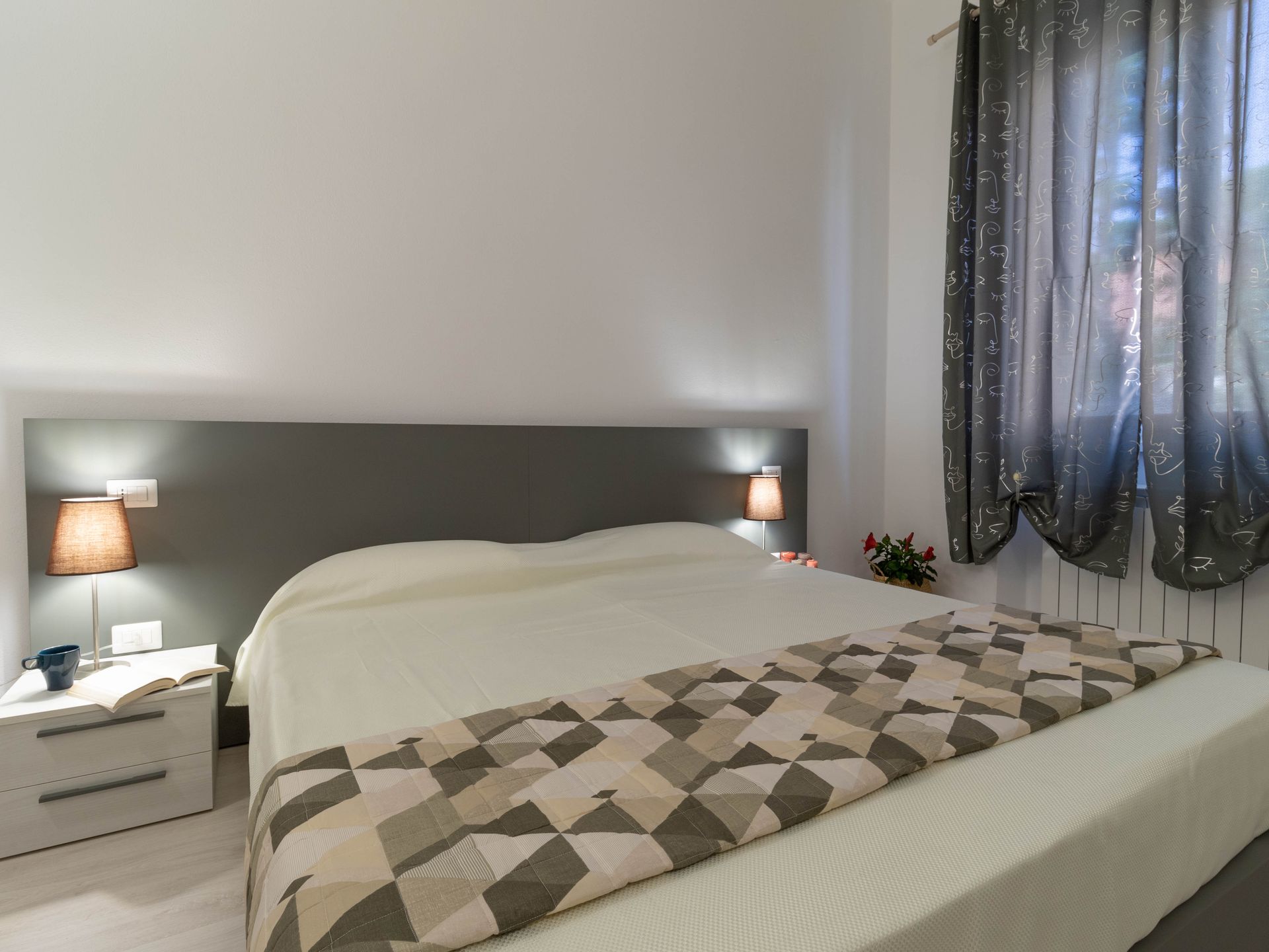 Camera da letto minimalista con un grande letto, testiera grigia, lampade da comodino e tende trasparenti alla finestra.