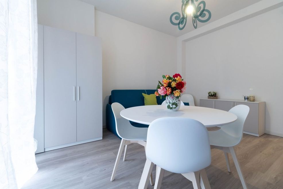 Luminosa sala da pranzo con tavolo bianco, sedie blu, armadio e fiori accanto a una finestra soleggiata.