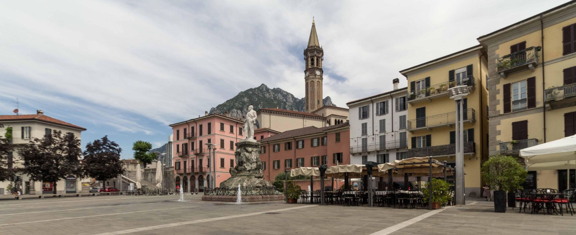 Piazza storica di una cittadina europea con edifici colorati, tavolini dei caffè e un alto campanile sotto un cielo nuvoloso.