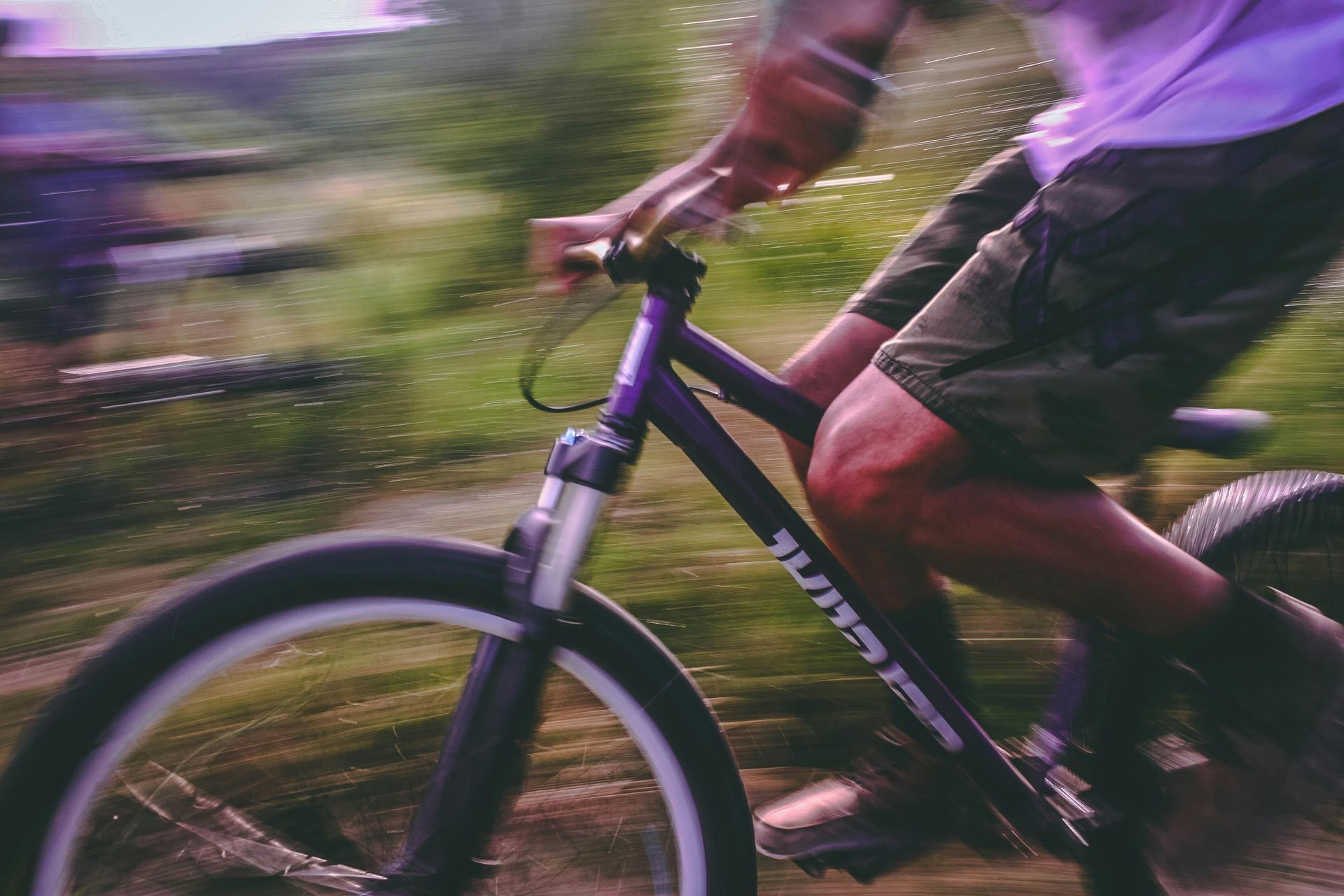 Ciclista di mountain bike in sella a una bici nera su un sentiero forestale sfocato, con indosso pantaloncini e una maglietta viola.