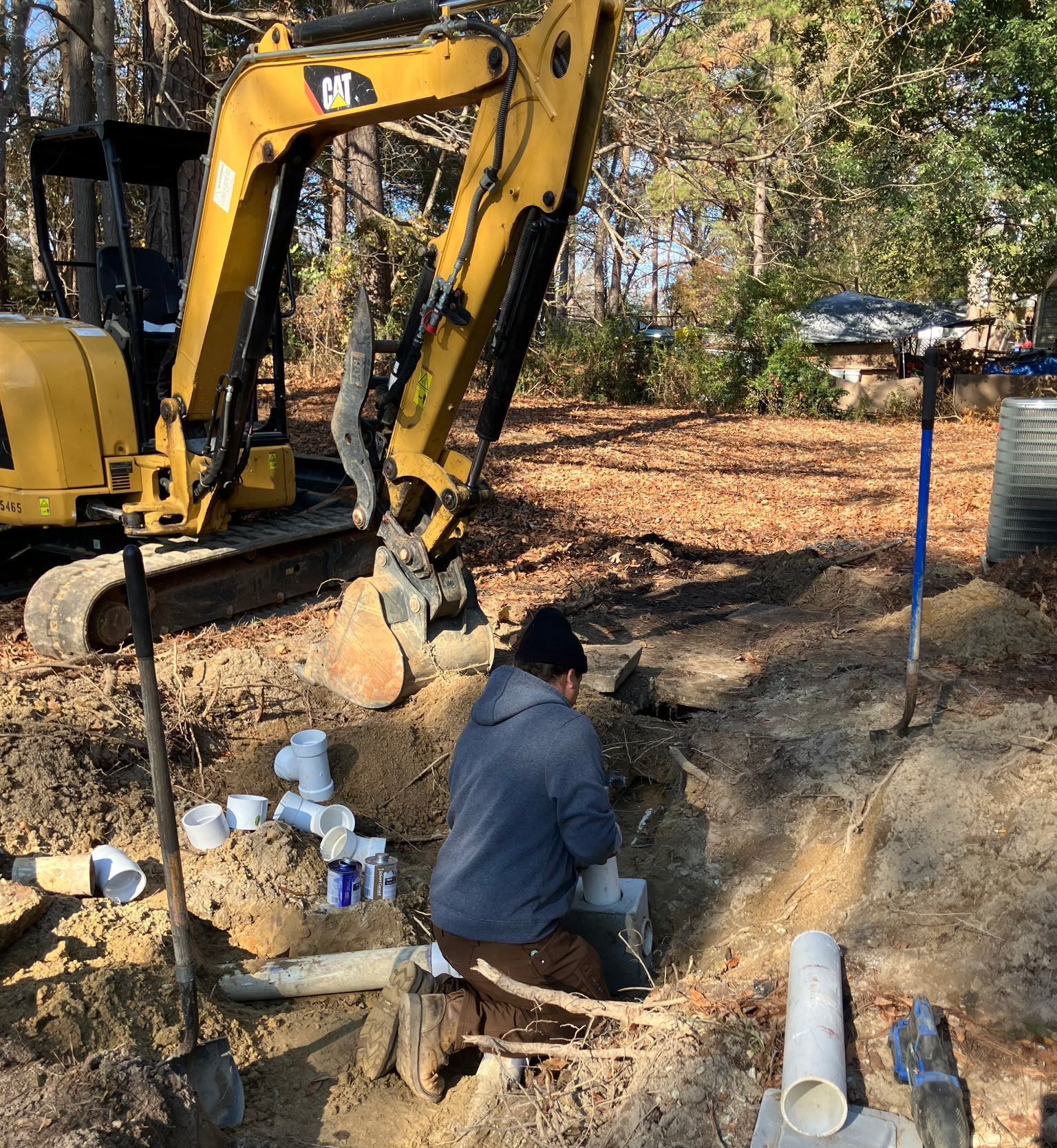 A person works in a trench with white pipes, an excavator in the background. Autumn leaves and trees.