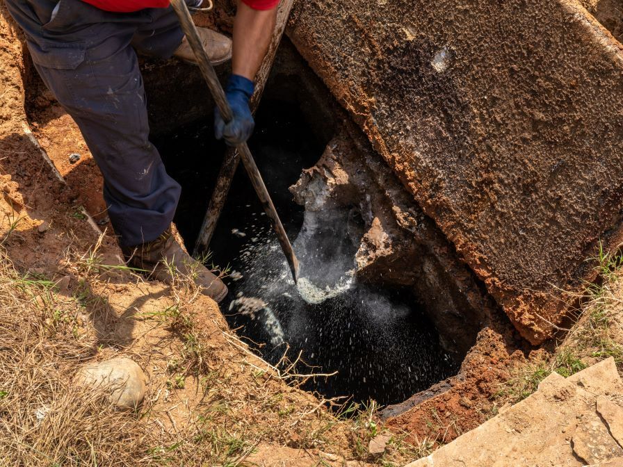 Person in blue gloves and pants cleaning a dark, open septic tank; brown soil and concrete frame in Lumberton, NC.