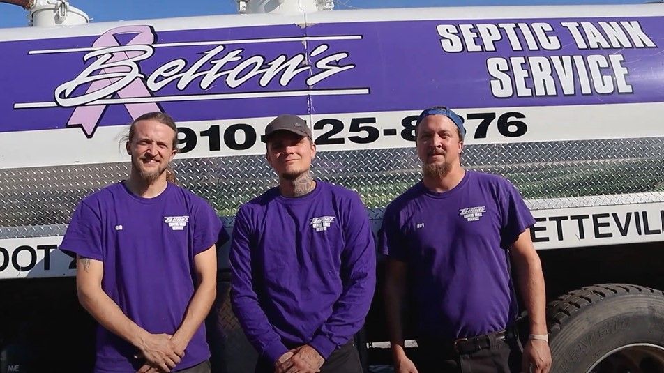 Three men in purple shirts stand in front of a septic tank truck with the company name, Belton's, and phone number in Lumberton, NC.