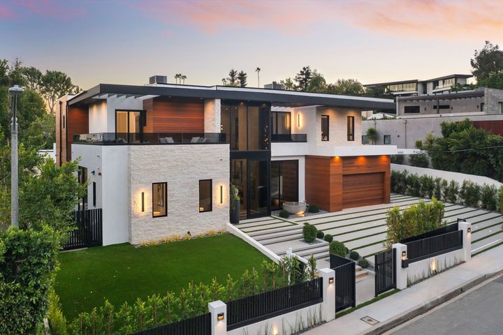 Modern two-story house with stone facade, warm lighting, manicured lawn, and a gated driveway at dusk