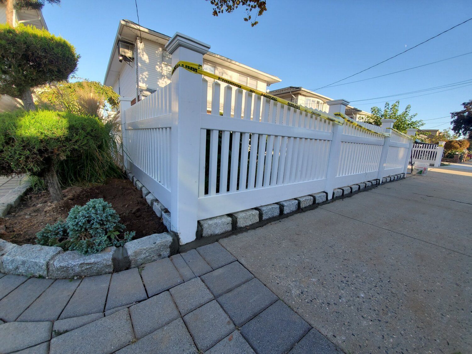 A white fence is surrounding a house on a brick sidewalk.