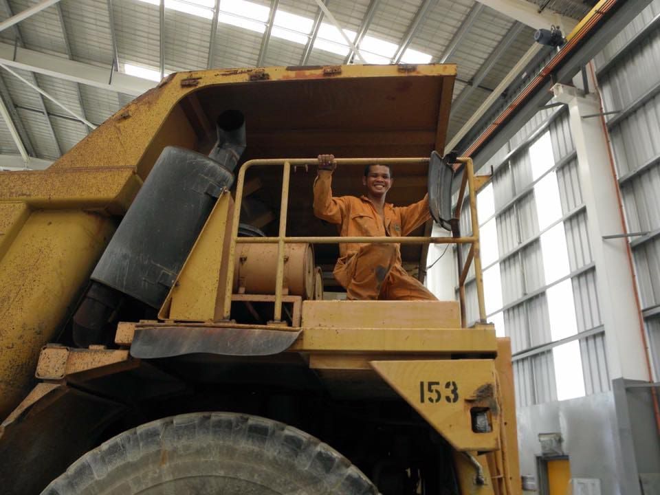 Man in orange jumpsuit smiles, seated in the cab of a large yellow mining truck. Industrial setting.