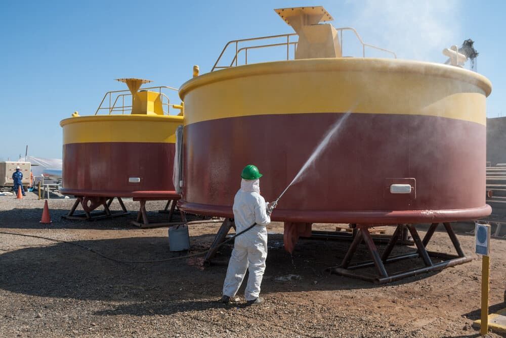 A Man in a White Suit is Cleaning a Large Tank With a High Pressure Washer — Waters Abrasive Blasting Services In Shaw, QLD