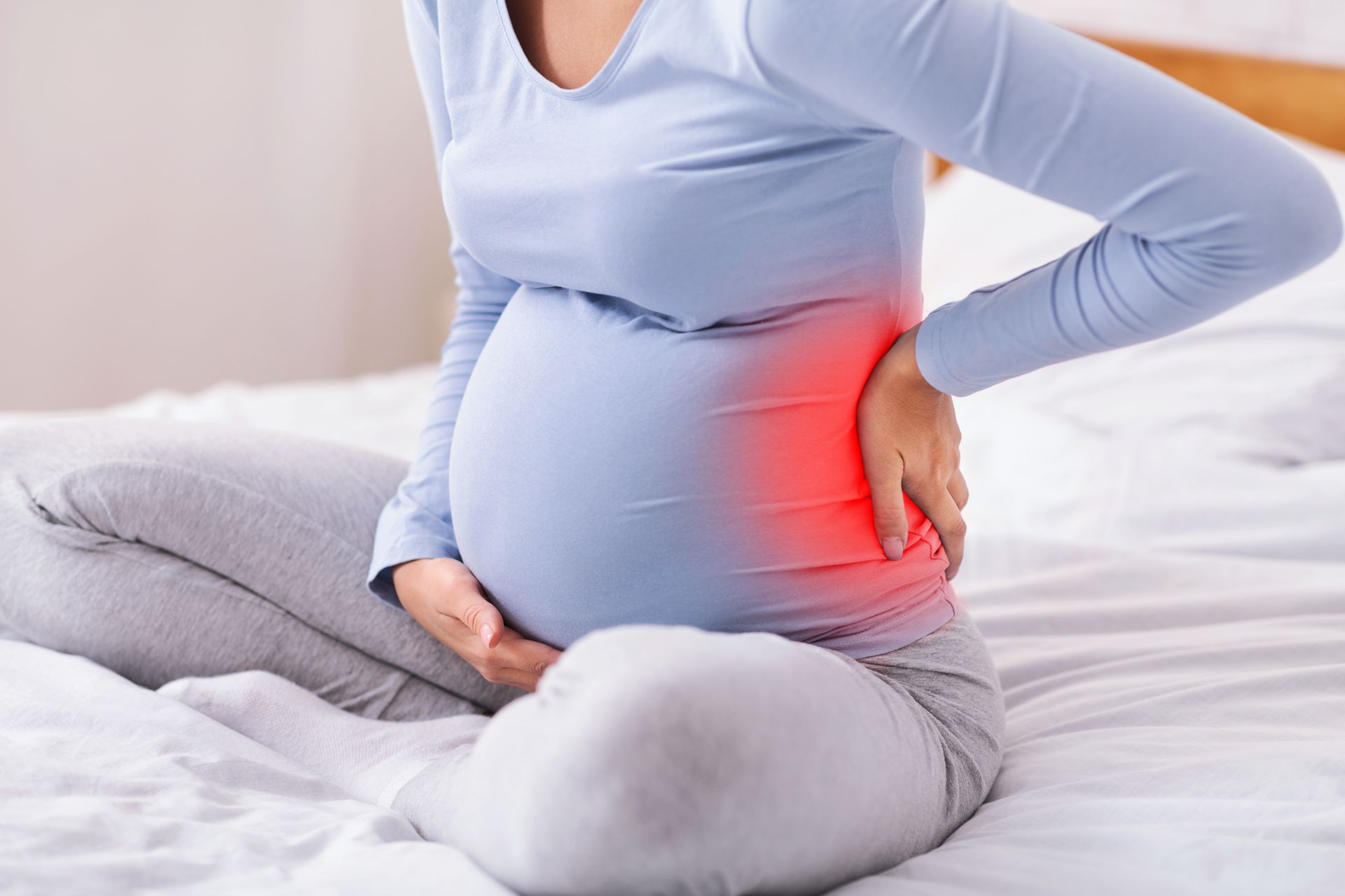 A pregnant woman is sitting on a bed holding her back in pain.