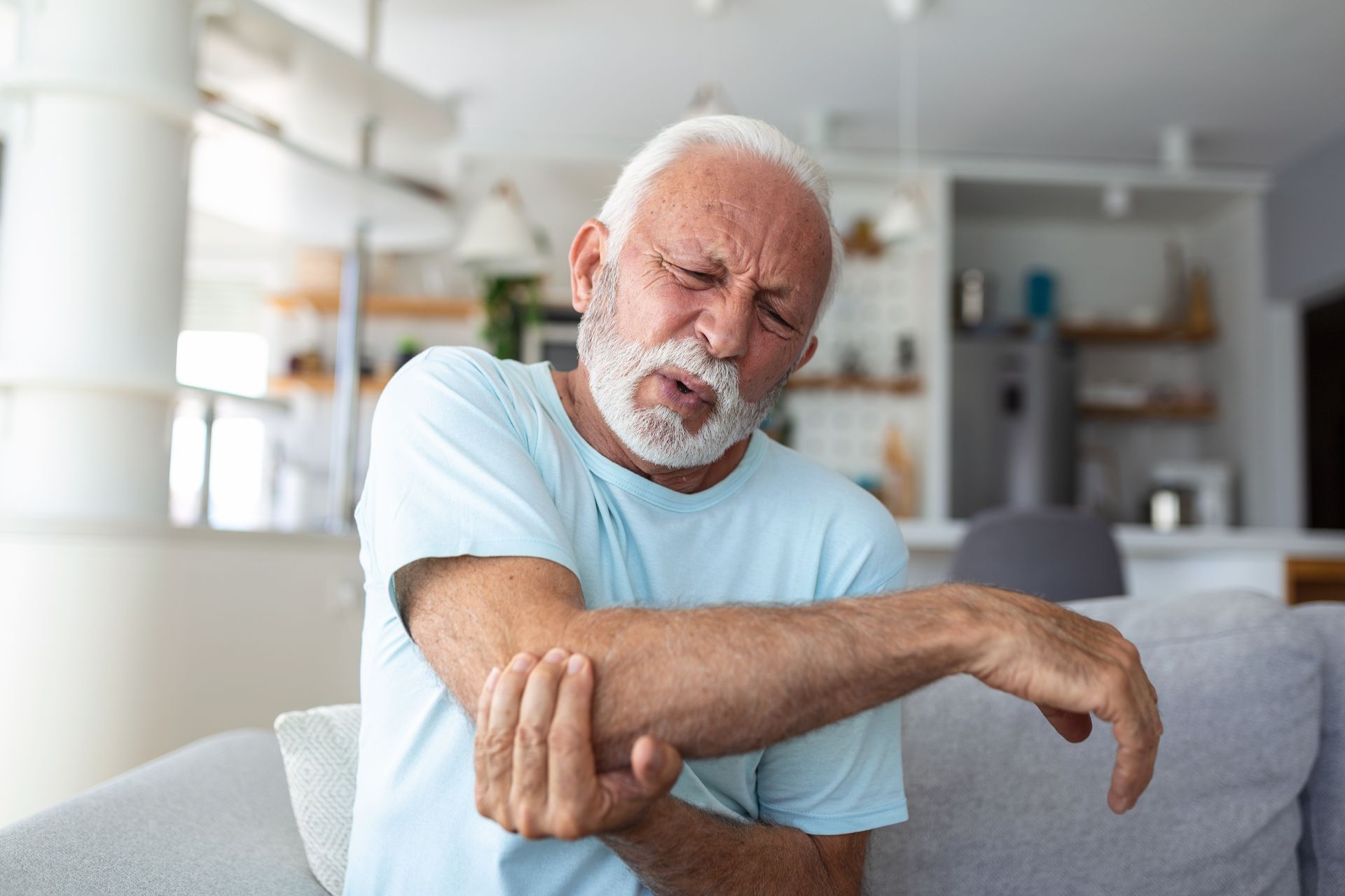 An elderly man is sitting on a couch holding his elbow in pain.