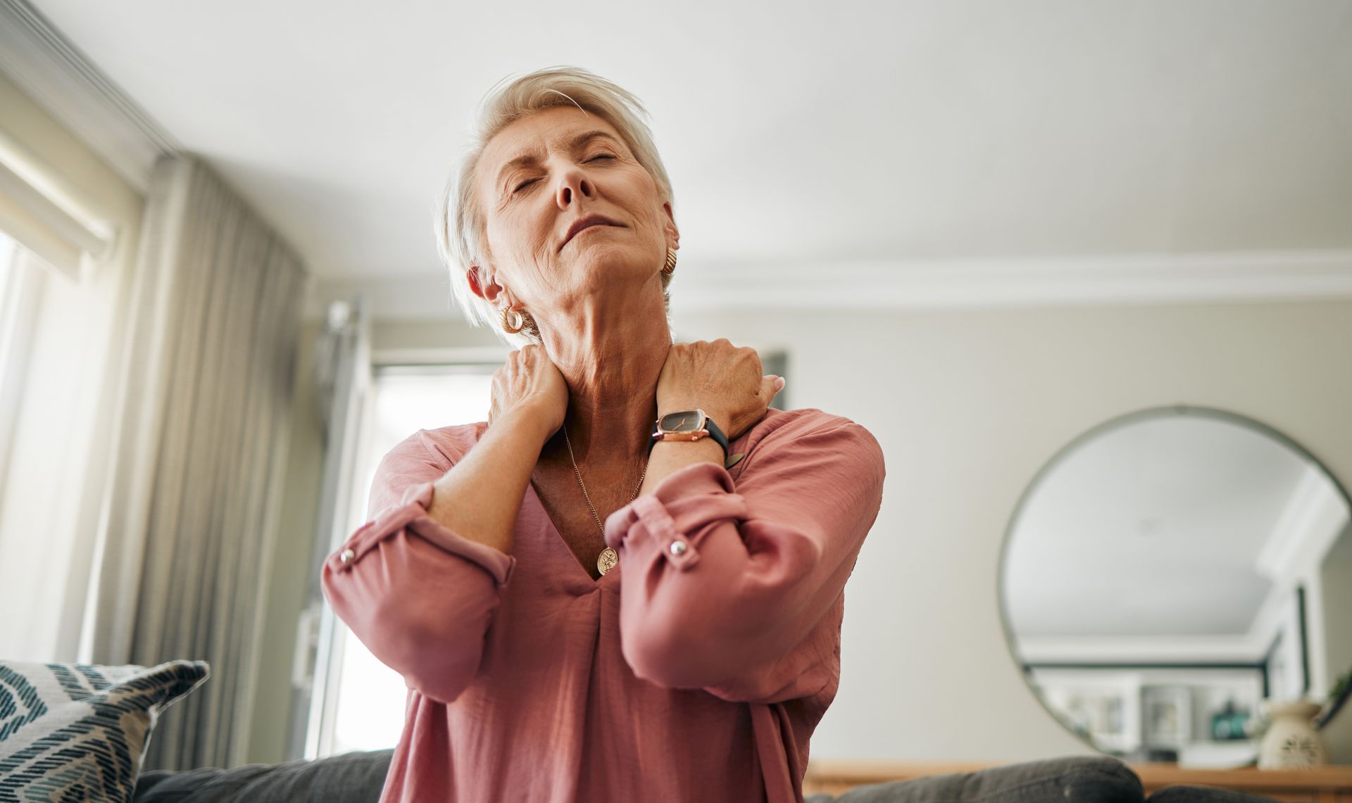 An elderly woman is sitting on a couch holding her neck in pain.