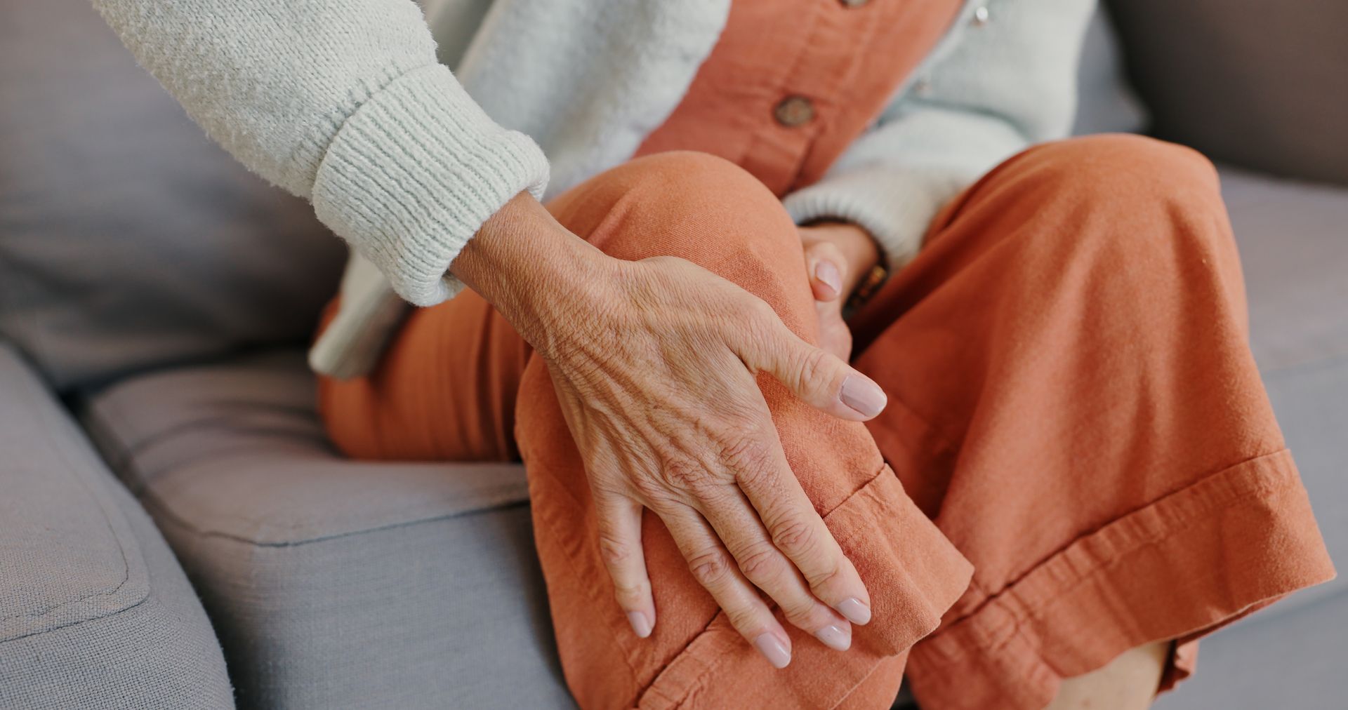 An elderly woman is sitting on a couch with her hands on her knees.