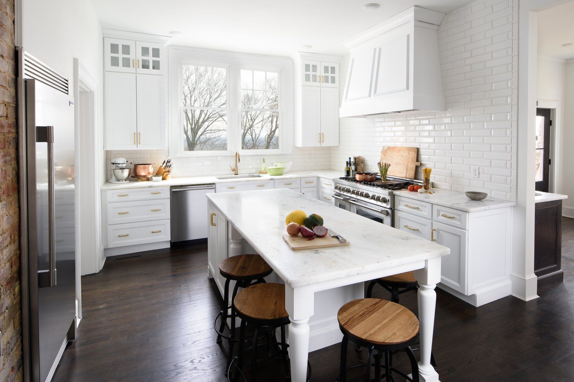 White kitchen with island, stove, stools, and a stainless steel fridge against a brick wall.