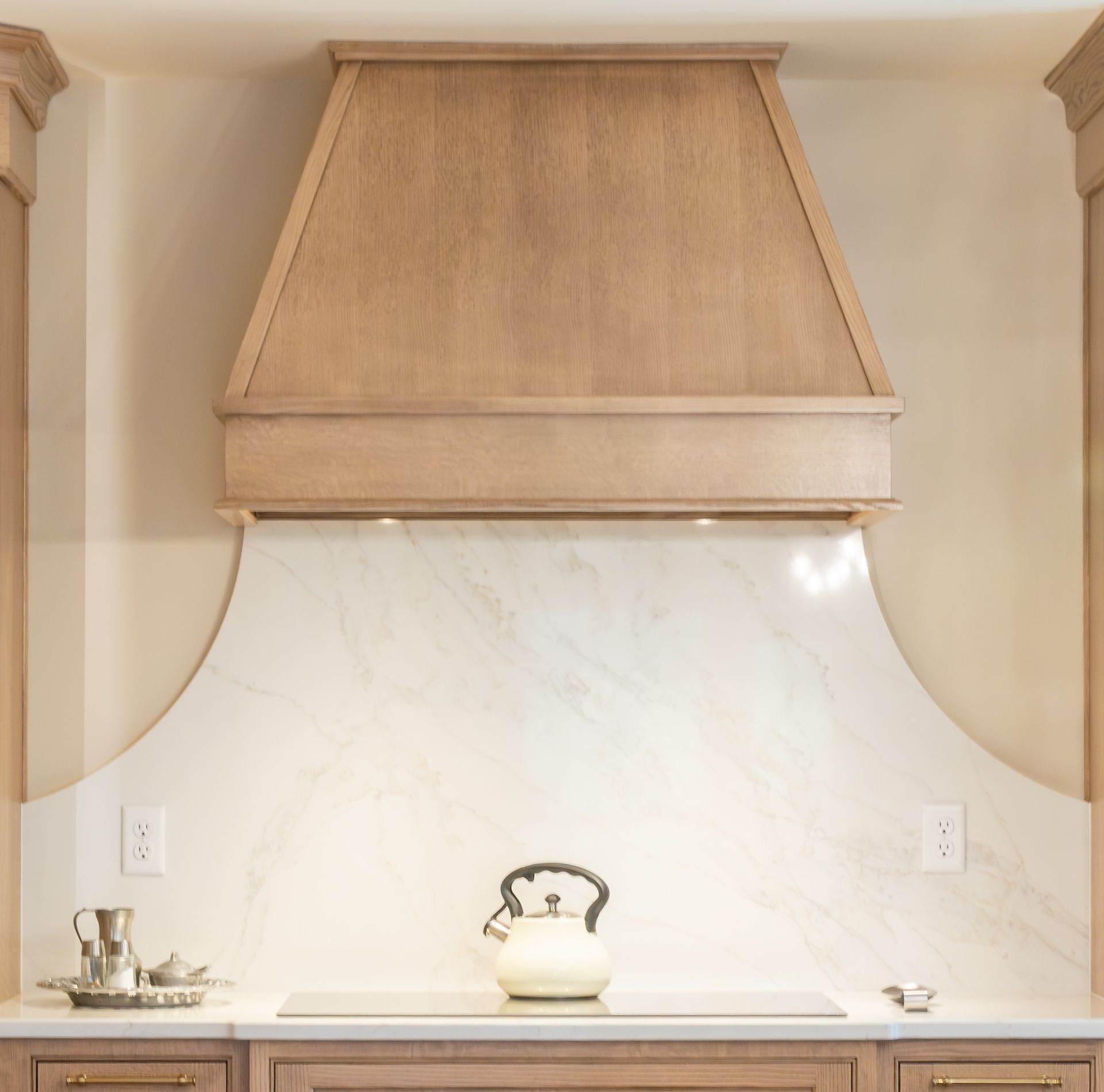 Beige kitchen range hood over a white marble backsplash and light wood cabinets