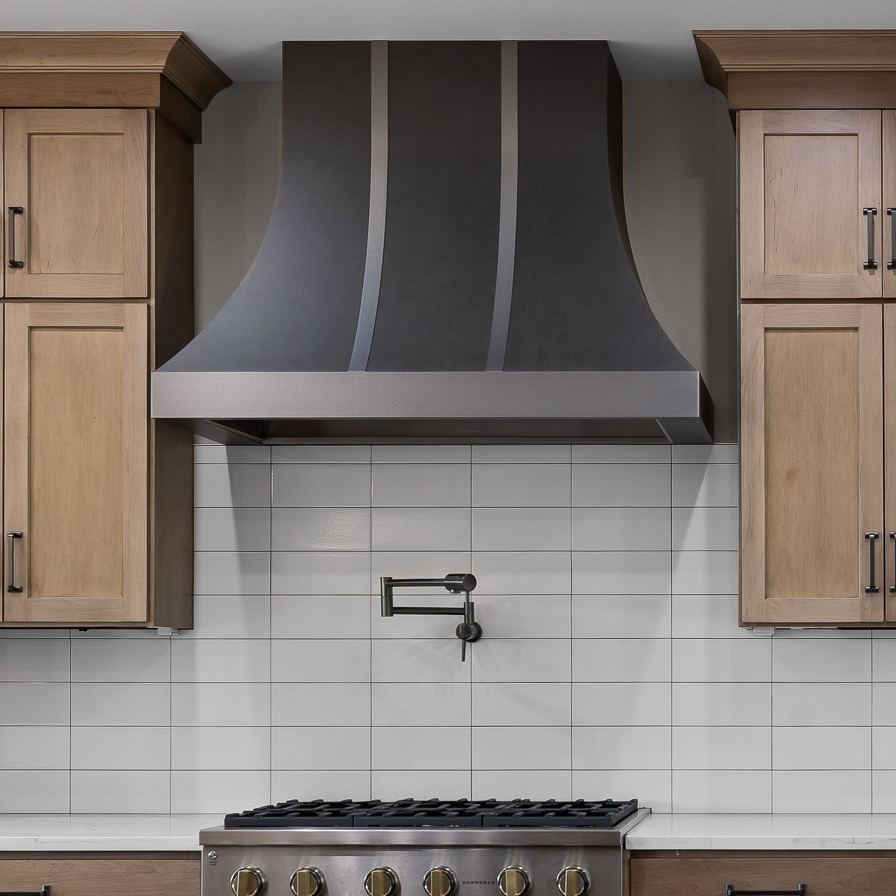 Modern kitchen with gray range hood, white tile backsplash, wood cabinets, and stainless stove