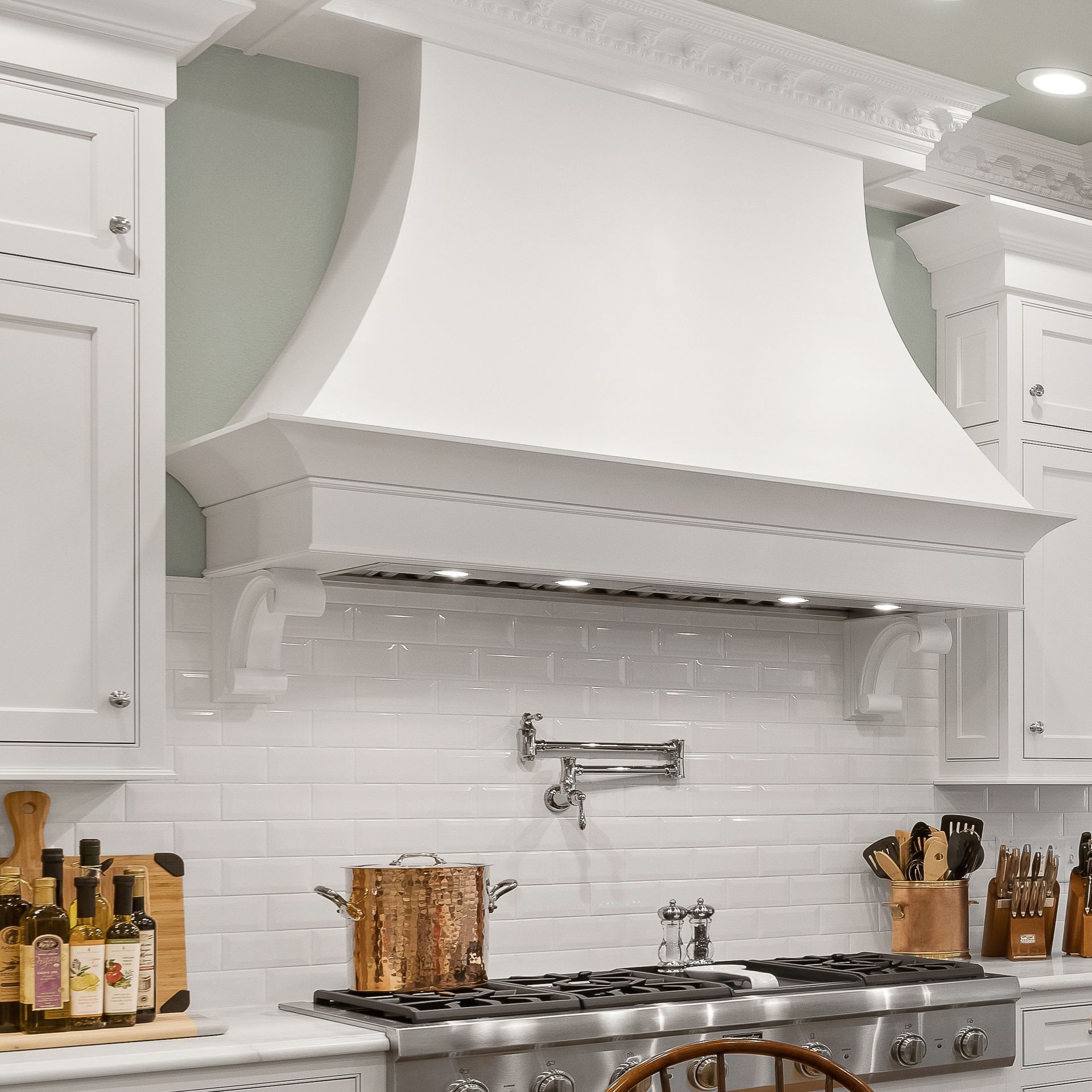 White kitchen with a large decorative range hood over a stainless steel stove and white subway tile backsplash