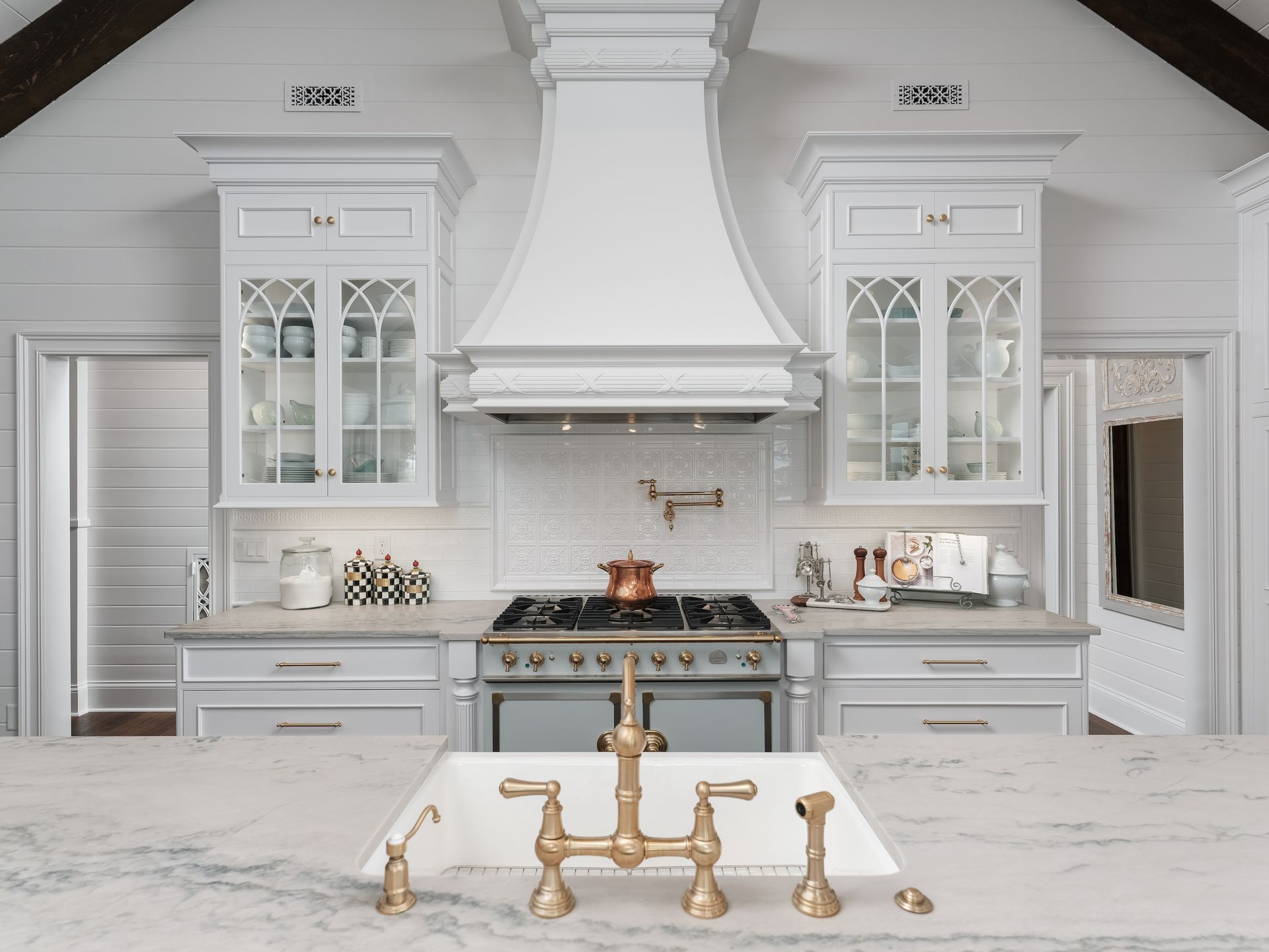 Elegant white kitchen with custom cabinets, stove, copper pot, and gold faucet.