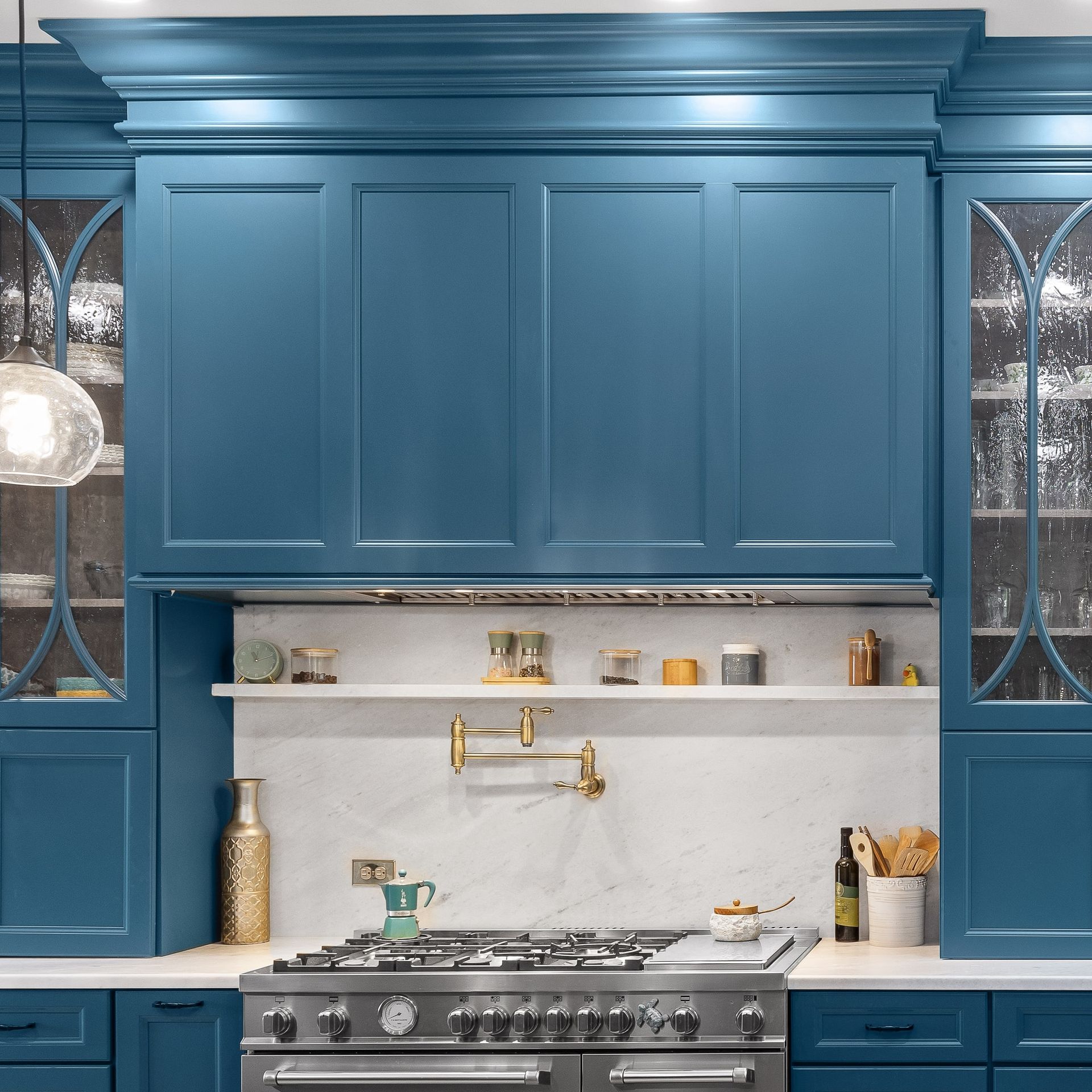 Blue kitchen with marble backsplash, brass faucet, and stainless steel stove beneath tall cabinets