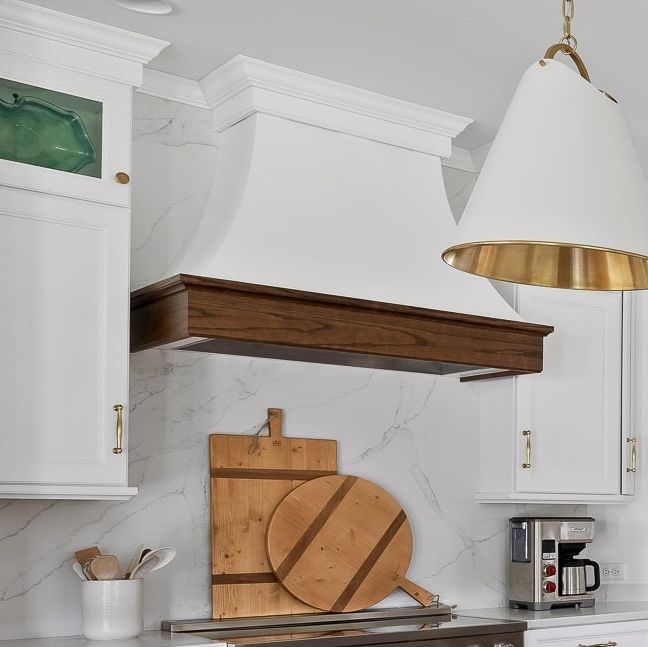 Bright white kitchen with wooden range hood, marble backsplash, hanging light, and cutting boards on the counter