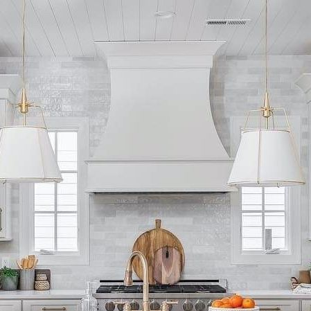 Bright white kitchen with a large hood, brass pendants, and a wooden cutting board by a sink.