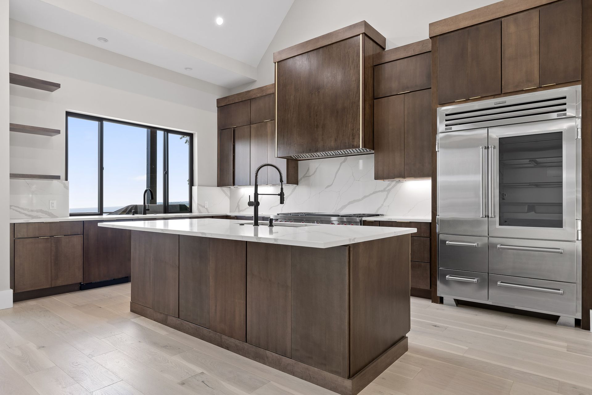 Modern kitchen with dark wood cabinets, white countertops, stainless steel refrigerator, and large window.