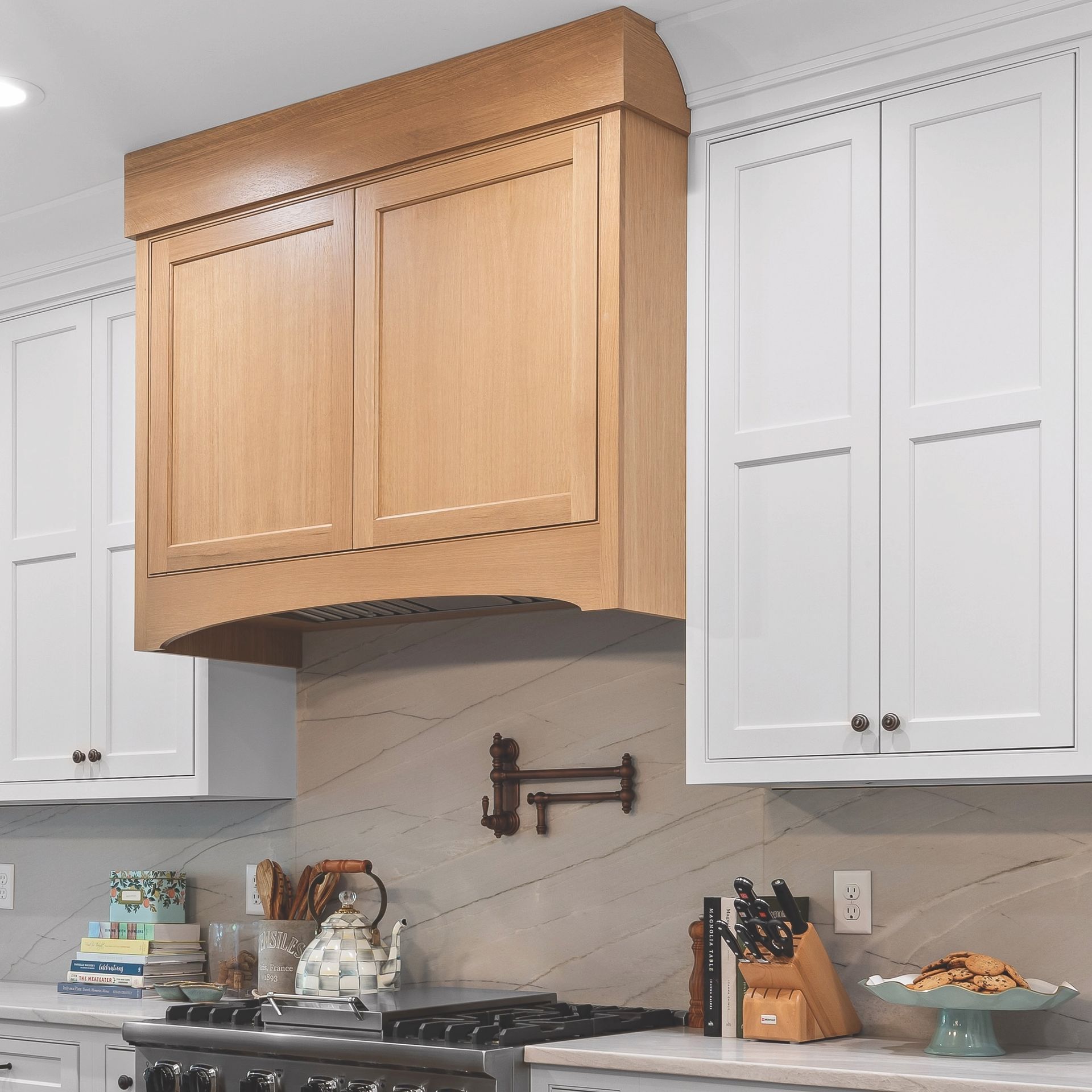 Modern kitchen with a large wood range hood over a stove, flanked by white cabinets and marble backsplash.