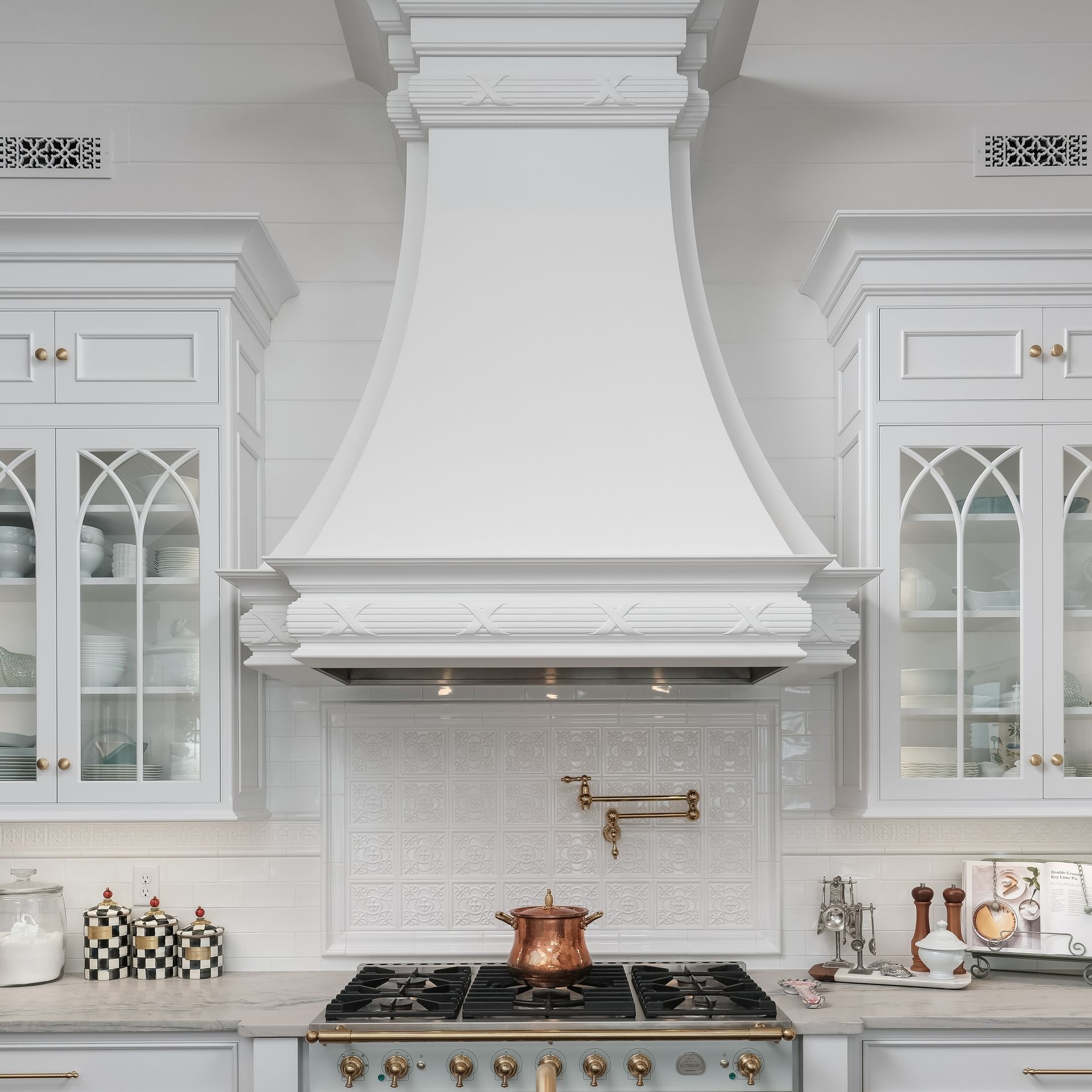 Bright white kitchen with ornate range hood, glass cabinets, and a copper pot on the stove