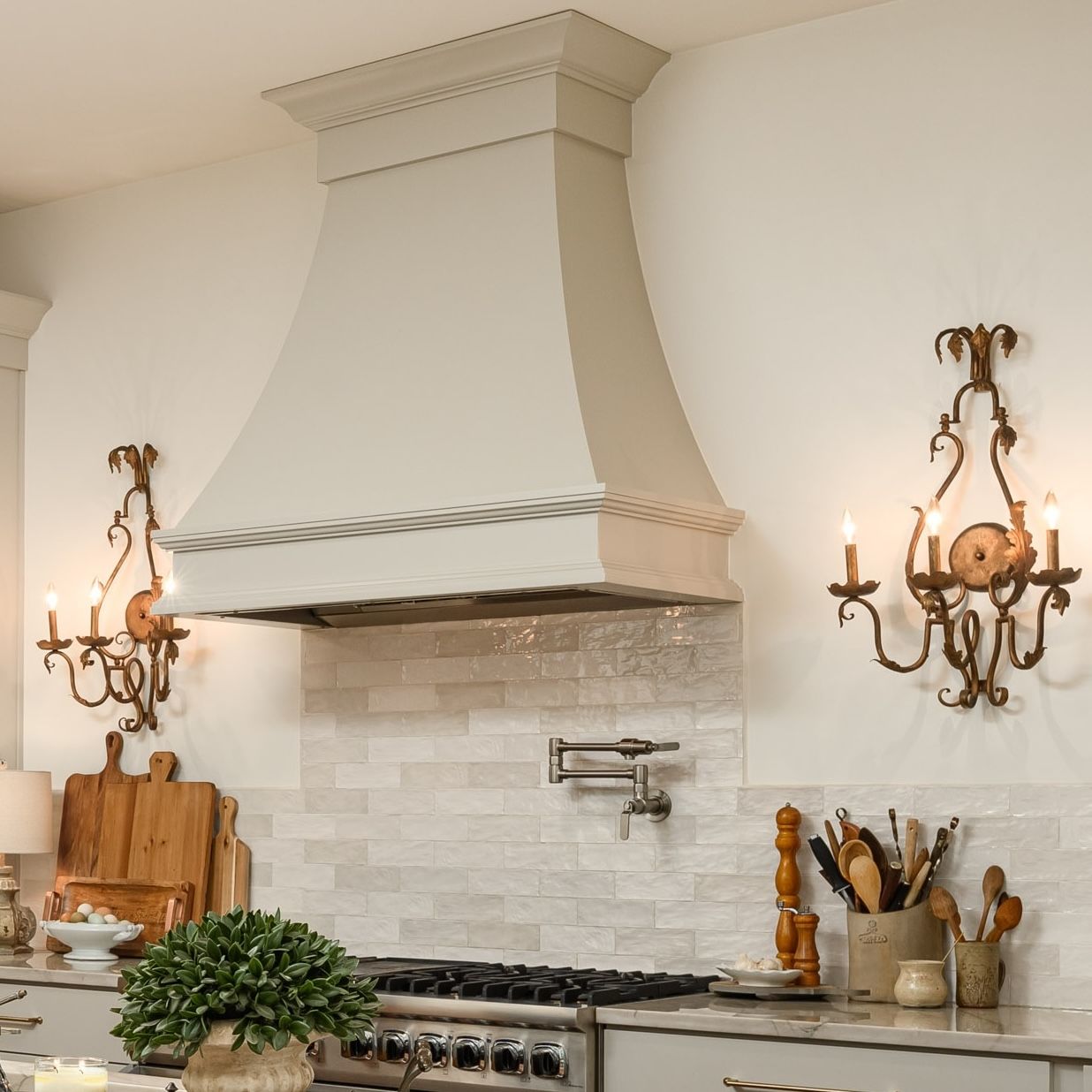Kitchen with white range hood, tiled backsplash, lit wall sconces, and countertop utensils