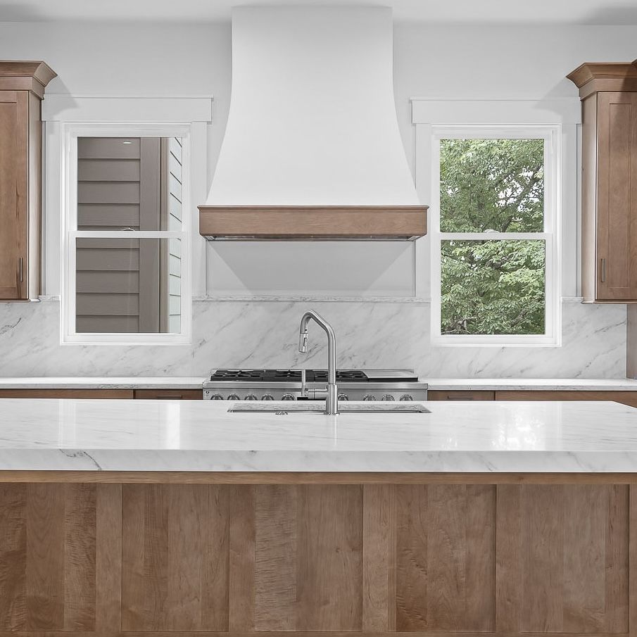 Modern kitchen with white marble island, wood cabinets, stainless range hood, and two windows over the sink