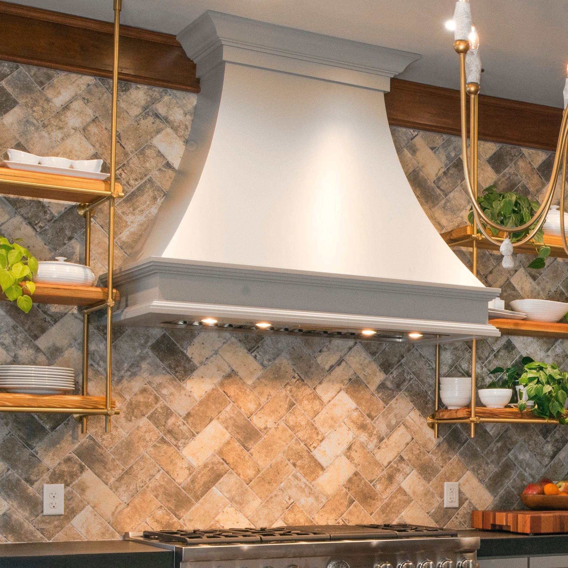 Modern kitchen with white range hood, stone backsplash, and brass shelves with plants and decor
