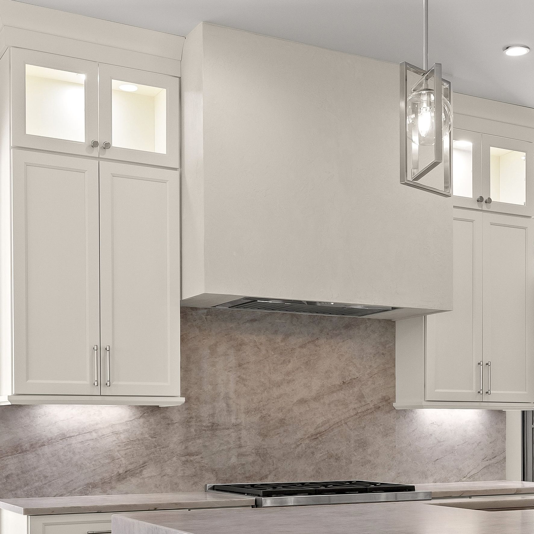 Modern white kitchen with upper cabinets, marble backsplash, and stainless range hood over a cooktop