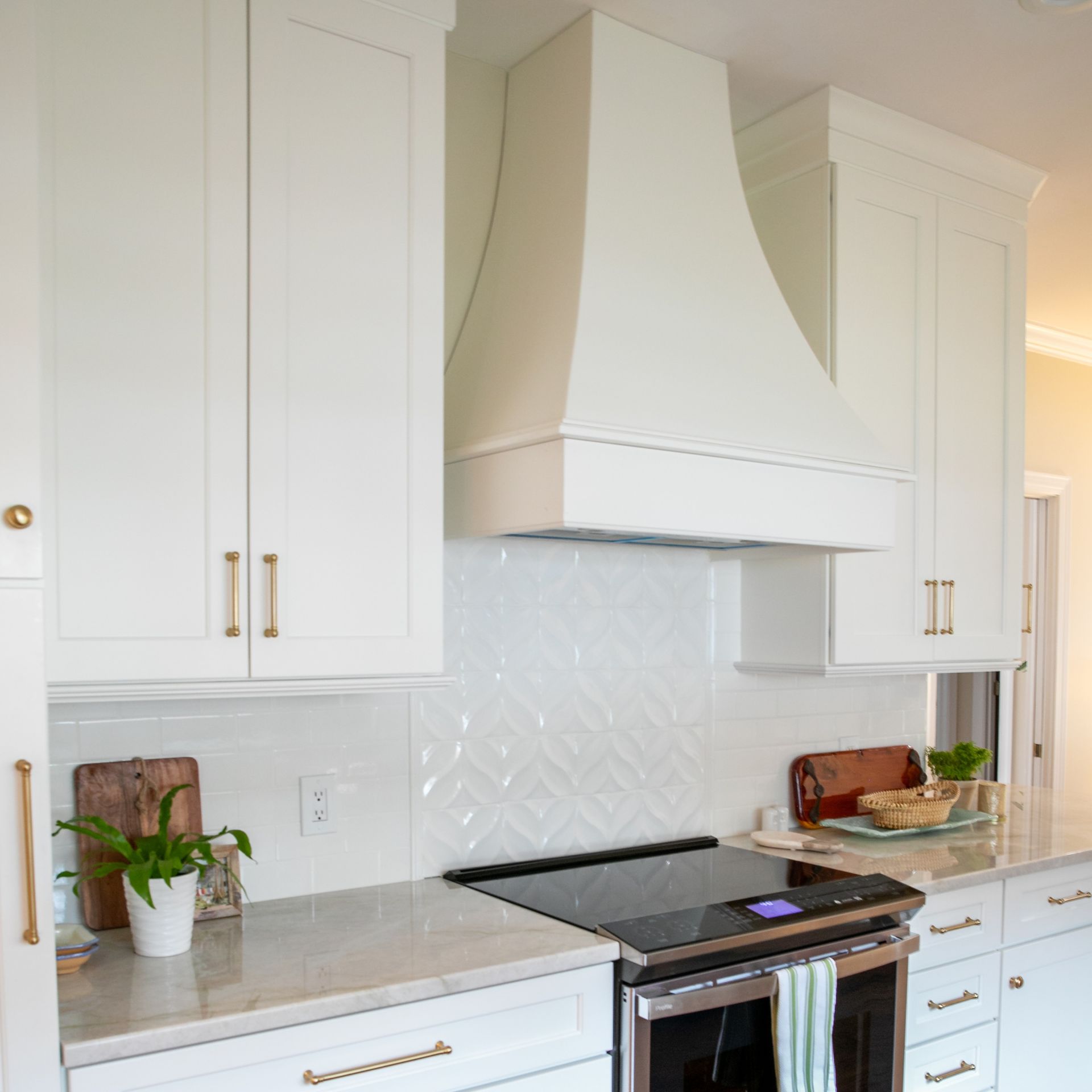 Bright white kitchen with brass handles, marble countertops, and a black stove beneath a white range hood