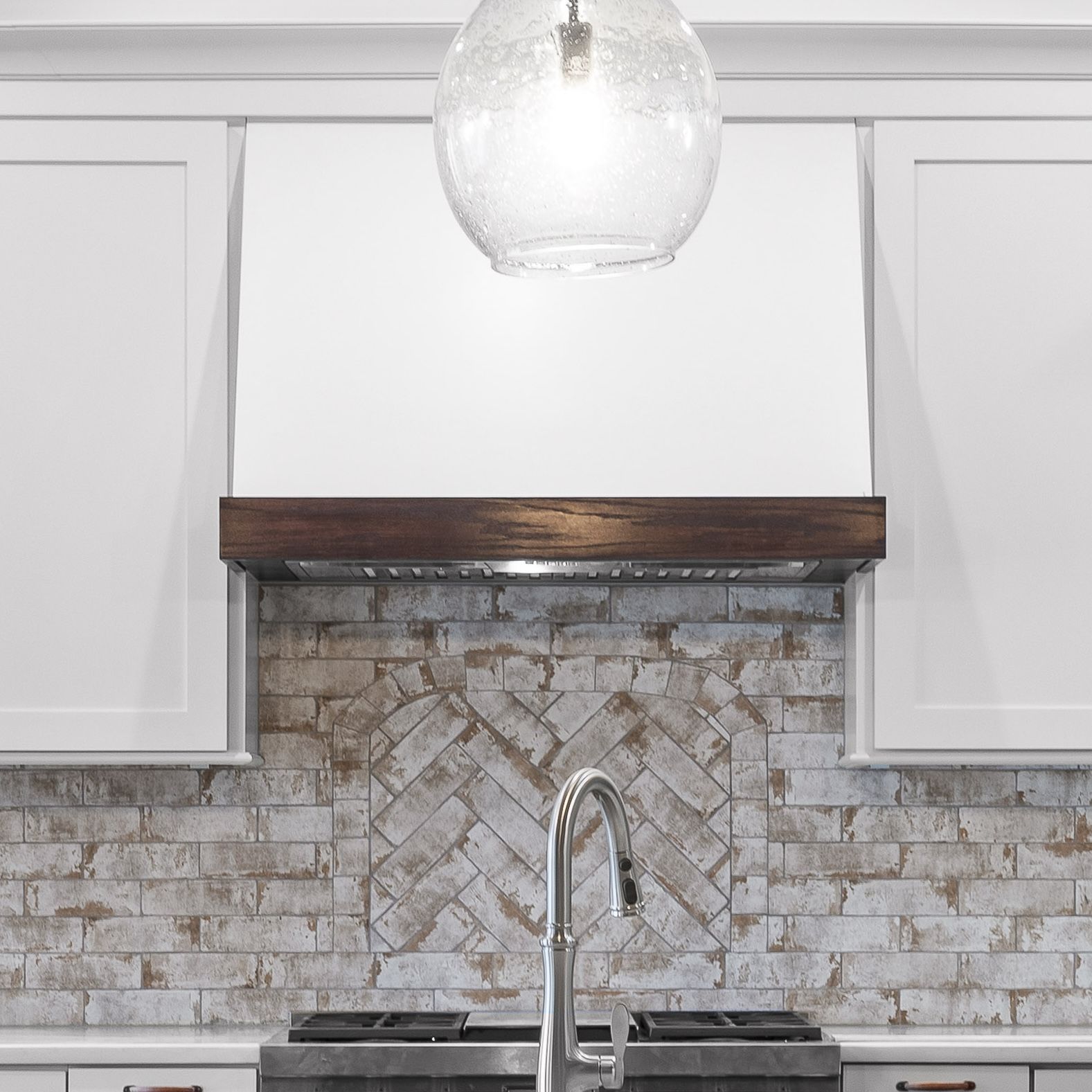 Modern kitchen with white cabinets, gray brick backsplash, wooden range hood trim, and glass pendant light