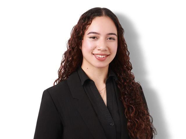 Smiling person with long curly hair wearing a black blazer on a white background