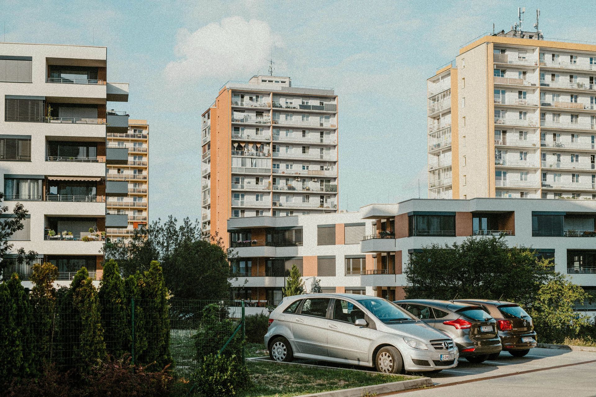 Apartment buildings and parked cars beside a grassy lot under a clear sky.