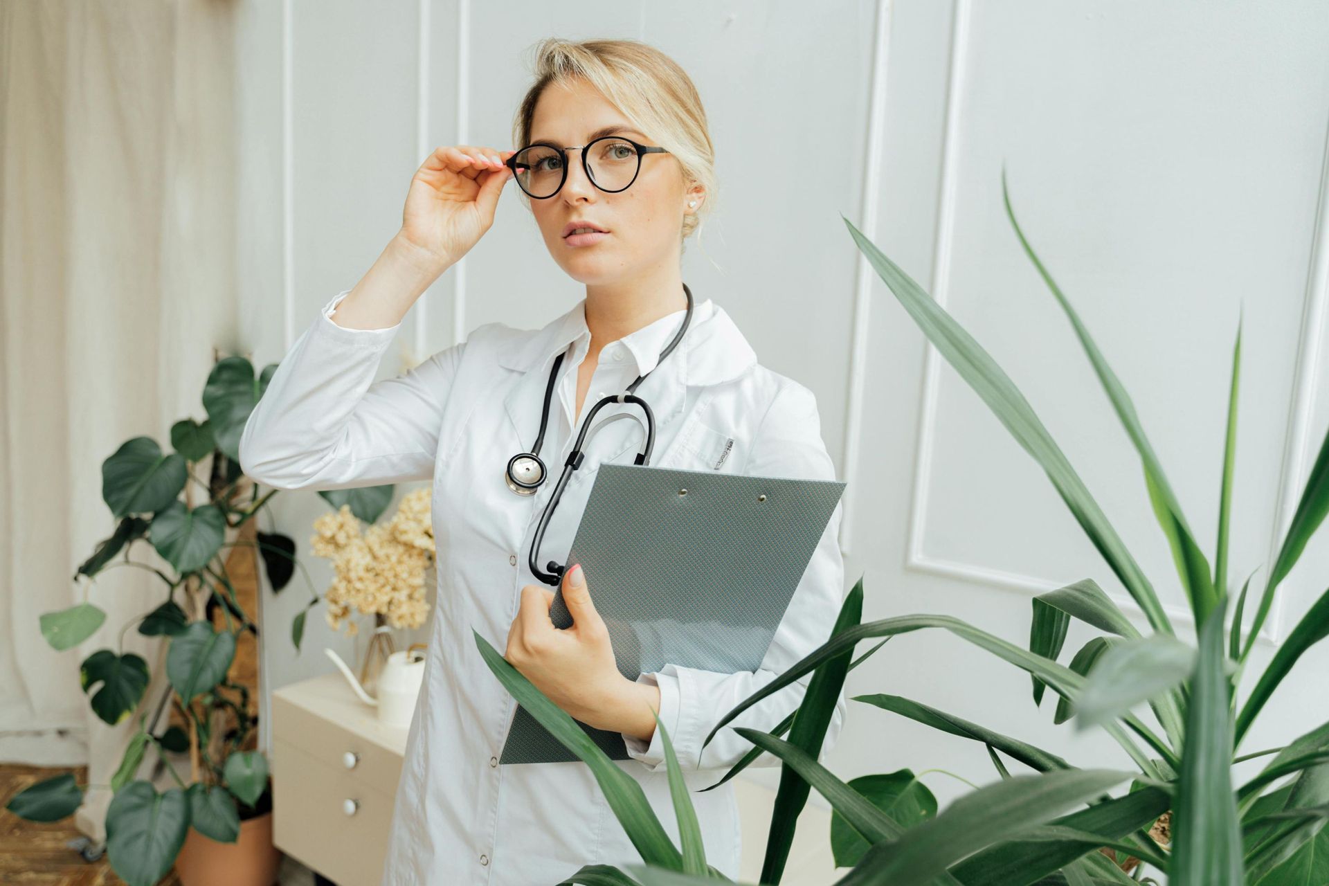 Doctor holding a clipboard in a bright clinic, adjusting glasses beside leafy plants