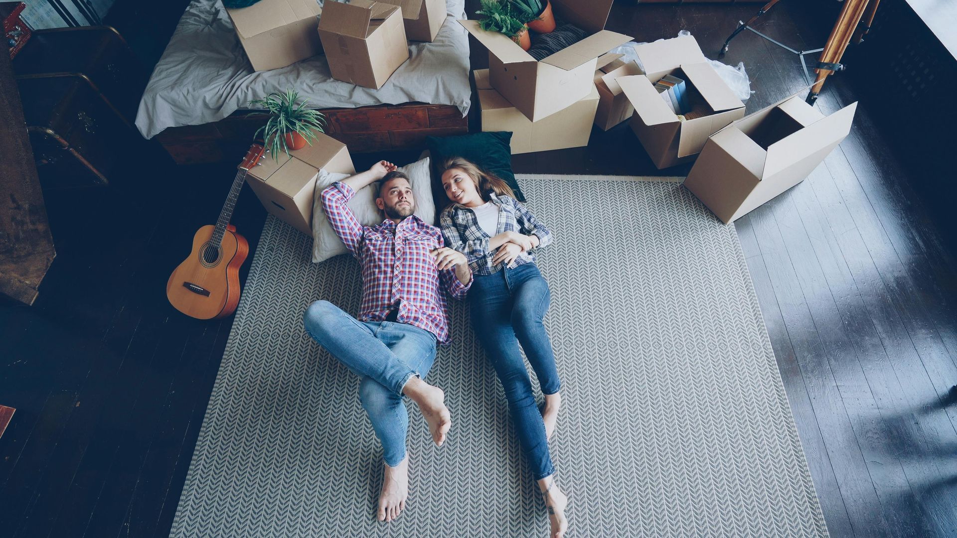 Two people lying on a rug among moving boxes, looking up in a room with a guitar and packing paper.
